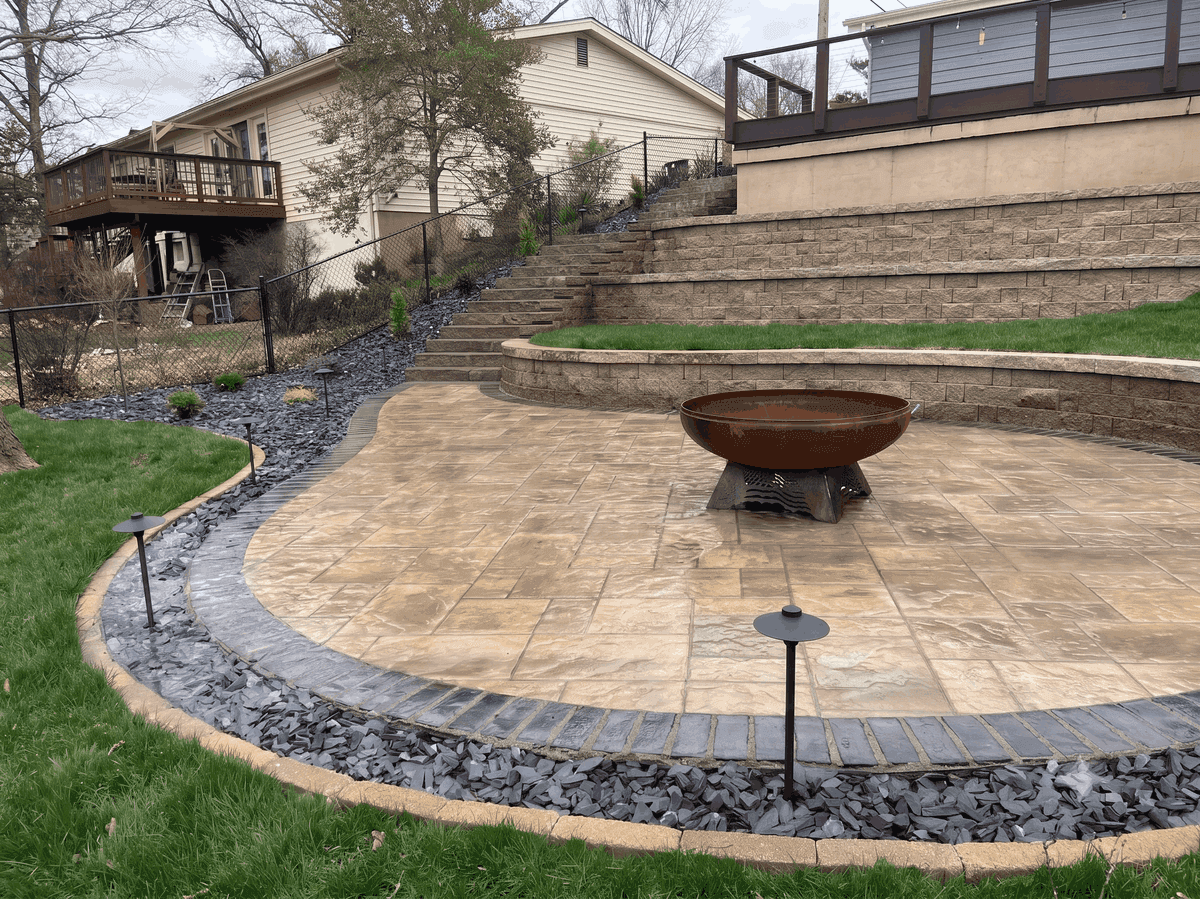 multi-level paver patio in a tiered backyard. The patio features distinct paving stones, a dark slate rock border, integrated path lighting, and a large, weathered steel fire bowl on a decorative base
